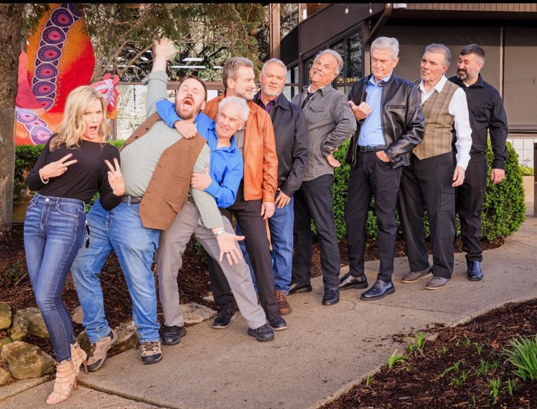 Group of adults posing playfully outside a building, with one man carried and others striking poses to the side.