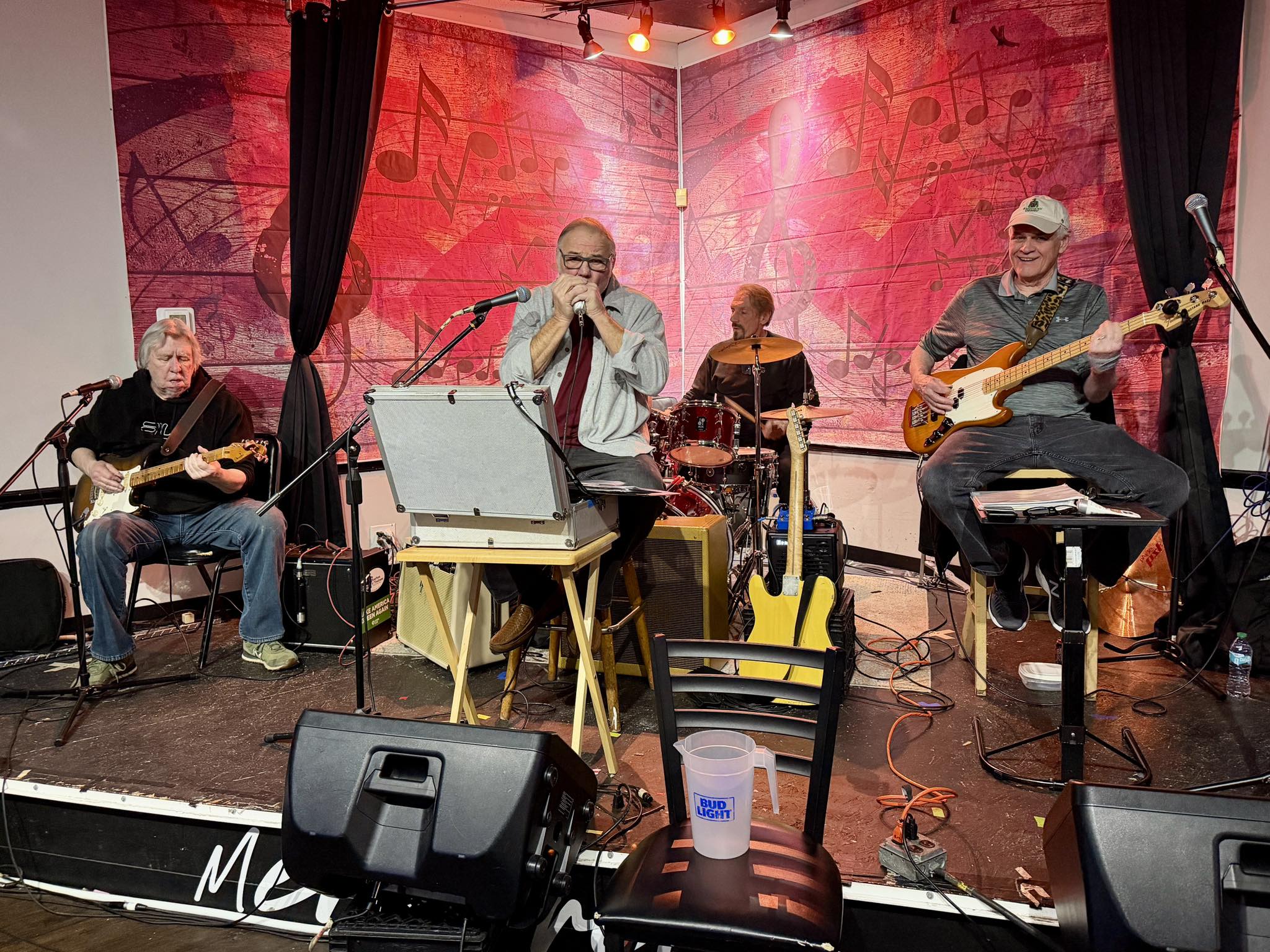 The Rhythm Cats - Turtle Run Winery Band playing on a small stage: guitarist left, vocalist center, drummer behind, bassist right; red backdrop with musical notes and stage gear in view.