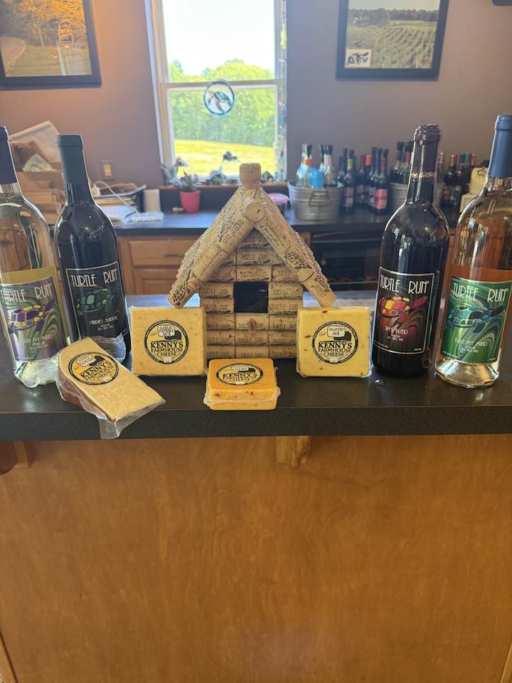 cheese lovers weekend - Turtle Run Winery Blocks of cheese and cheese-wrapped pieces sit on a bar counter around a small wooden house centerpiece; several wine bottles line the back, with a window and bottles visible in the background.
