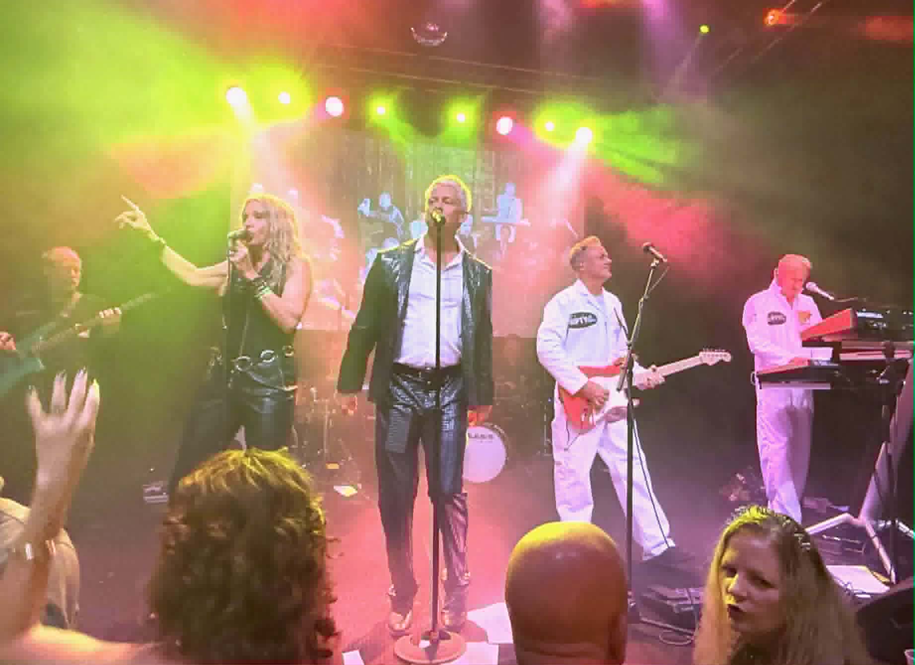 Live band performs on stage under colorful yellow, green, and pink lights, audience raising hands in front of them.