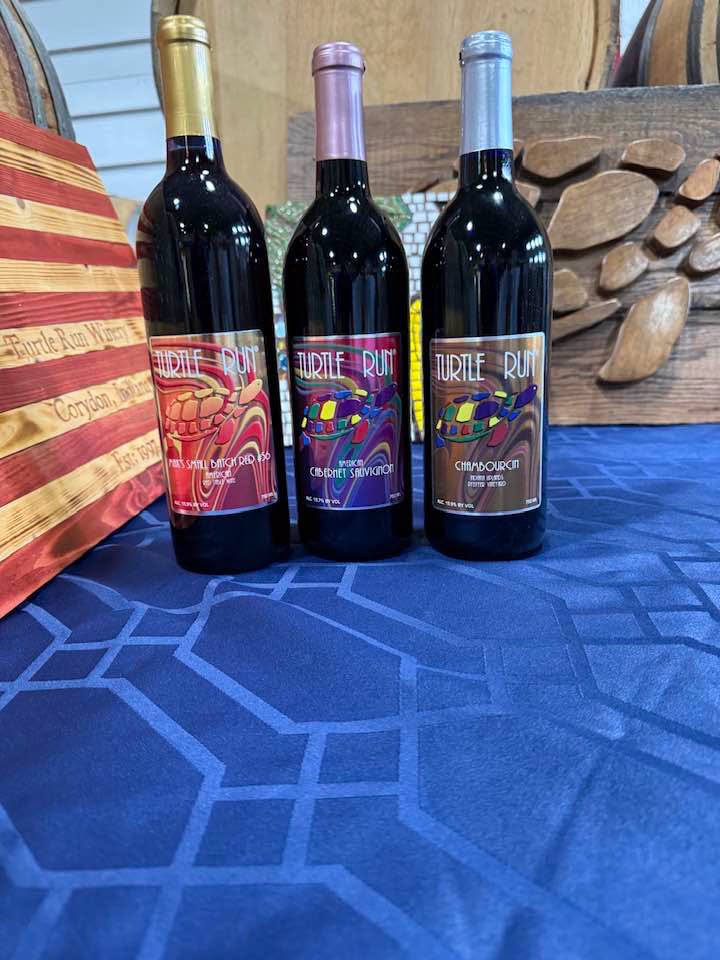 Three Turtle Run wine bottles with red, purple, and blue labels stand on a blue geometric tablecloth, with wooden barrels in the background.